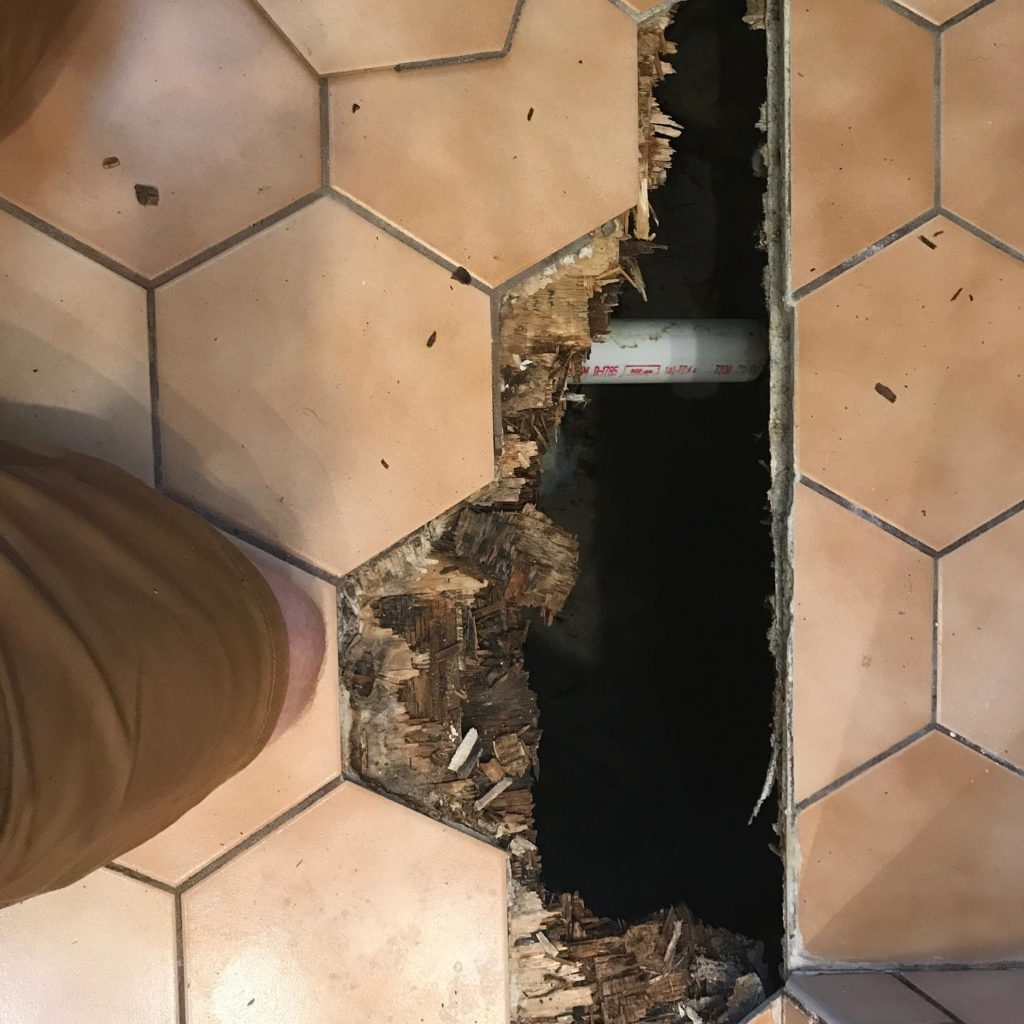 Damaged tile floor with a large hole exposing wood subfloor and a visible PVC pipe underneath.